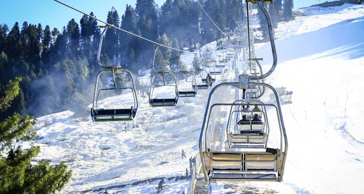 Télésiège montant au-dessus d'une pente de montagne enneigée avec des skieurs en contrebas et une forêt de pins environnante.