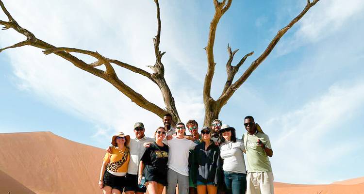 Un groupe de touristes pose sous un épineux-chameau mort et nu au milieu des dunes orange ondulantes de Sossusvlei.