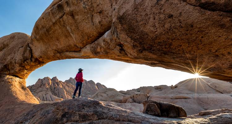 Persona parada sobre una formación rocosa con el sol asomándose a través del arco y montañas en el fondo.