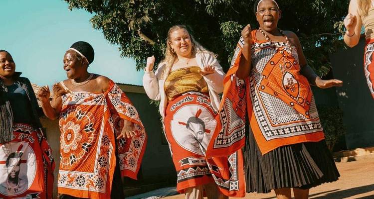 Vrouwen in kleurrijke traditionele Swazi-kleding dansen en zingen buiten onder een blauwe hemel.