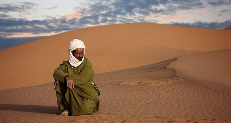Man gekleed in traditionele kledij zittend op woestijnzand.