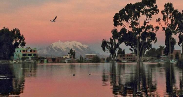 Un lago tranquilo al atardecer con reflejos de montañas y casas a lo largo de la orilla.