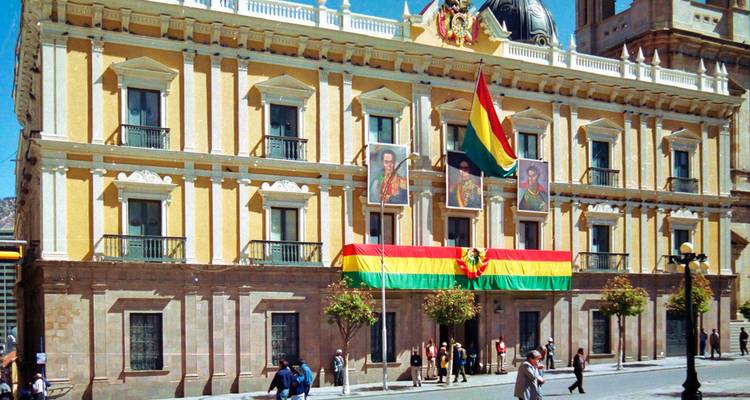 Un edificio gubernamental histórico con banderas en su fachada, personas caminando cerca en un día soleado.