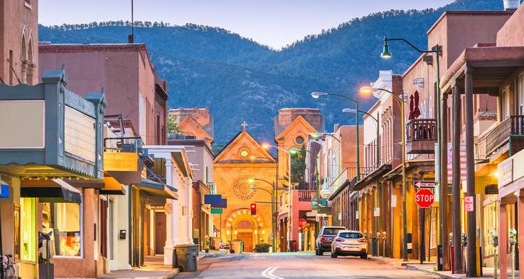 Historische Straße in Santa Fe, die zur beleuchteten Kathedralbasilika mit Bergkulisse in der Abenddämmerung führt.