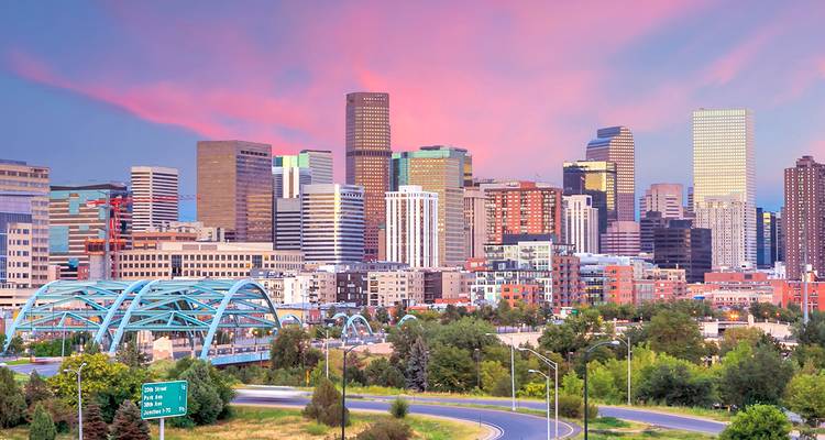 Lebendige Skyline von Denver bei Sonnenuntergang mit rosa Wolken über den Stadtgebäuden und einer blauen gewölbten Brücke.