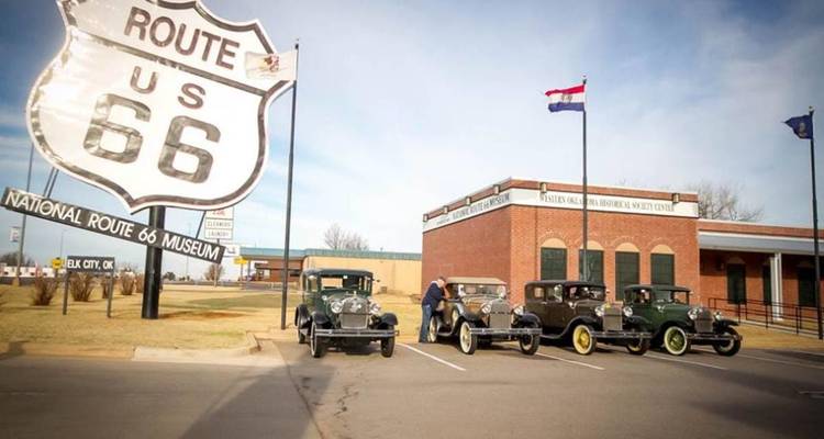 Exterior del Museo de la Ruta 66 Nacional con un enorme letrero de escudo y autos antiguos estacionados afuera bajo un cielo azul.