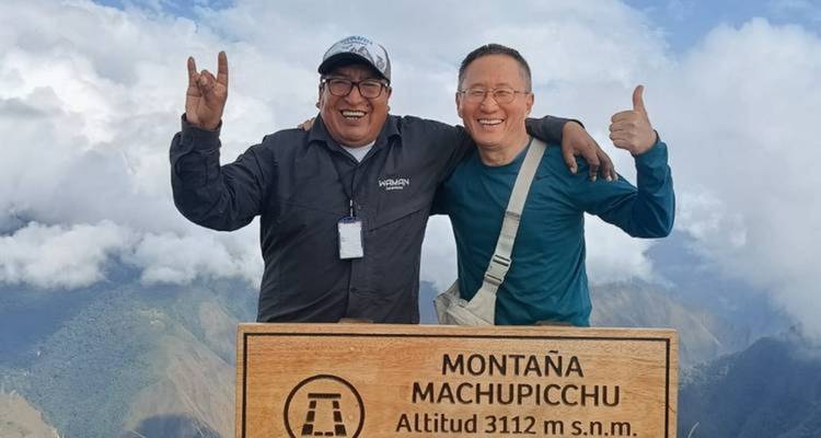 Dos trekkers sonrientes posando con un letrero de madera en la Montaña Machu Picchu muy por encima de los picos de los Andes cubiertos de nubes.