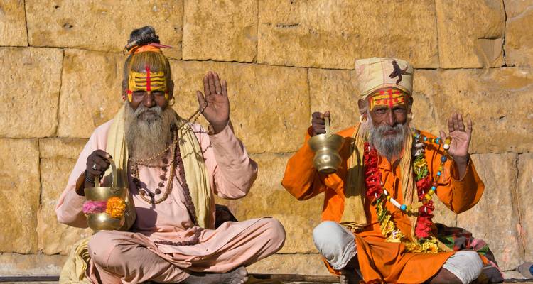 Dos sadhus vestidos coloridamente se sientan contra una pared de arenisca, bendiciendo la cámara con las manos alzadas.