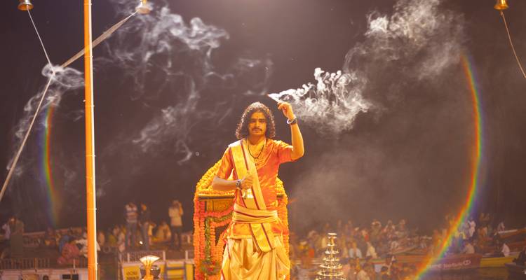El sacerdote realiza el aarti vespertino, elevando incienso con espirales de humo dramáticos contra un cielo oscuro y una multitud iluminada.
