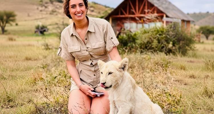 Voyageur souriant en tenue de safari kaki assis à côté d'un lionceau blanc joueur avec un lodge au toit de chaume en arrière-plan.