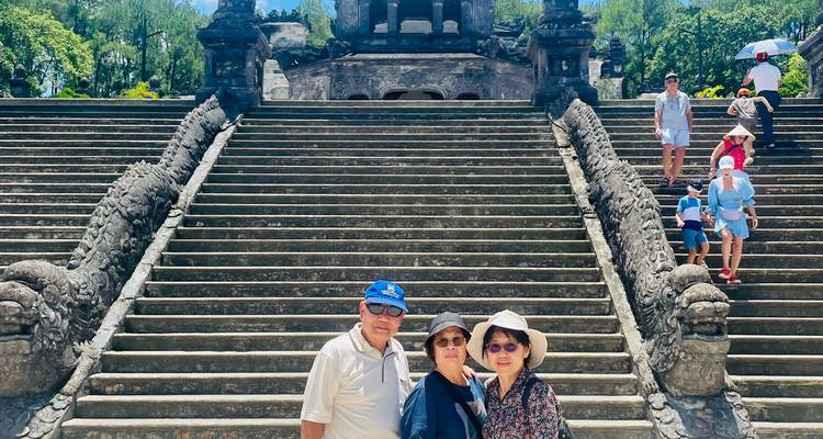 Trois voyageurs posant sur des escaliers monumentaux ensoleillés menant à une structure de tombeau ornée entourée de verdure.