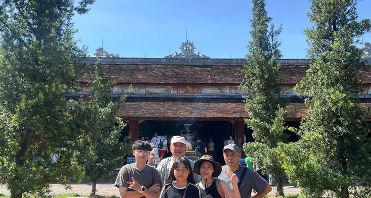 Cinq voyageurs posant entre de grands conifères devant un hall de temple vietnamien traditionnel.