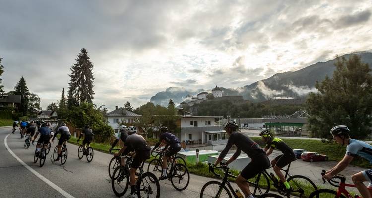 Un peloton de cyclistes sur route monte en serpentant une route de montagne sinueuse vers une forteresse enveloppée de brume.