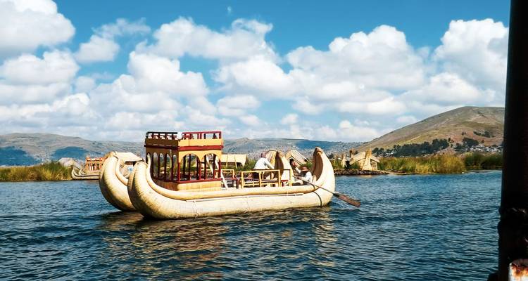 Un bateau traditionnel en roseaux avec auvent flotte sur les eaux bleu profond du lac Titicaca, encadré par des roseaux et des collines ondulantes sous des nuages blancs et cotonneux.