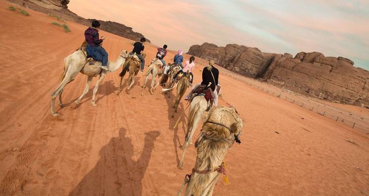 Caravan of riders on camels treks across red sand desert at sunset.