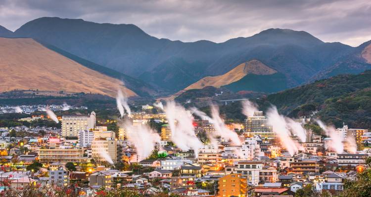Ville de Beppu avec de nombreuses colonnes de vapeur s'élevant sur un fond de montagnes au crépuscule.