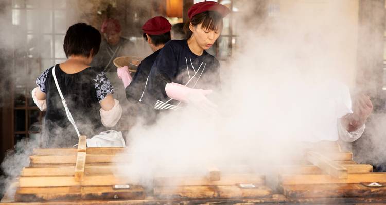 Les cuisiniers travaillent au-dessus de cuiseurs à vapeur en bois dégageant de la vapeur chaude dans une cuisine rustique.