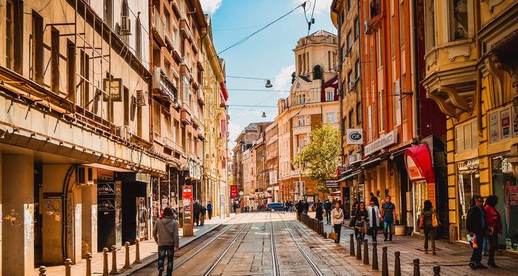 Lebendige Straße in Sofia mit Straßenbahngleisen, farbenfrohen Fassaden und Fußgängern unter blauem Himmel.