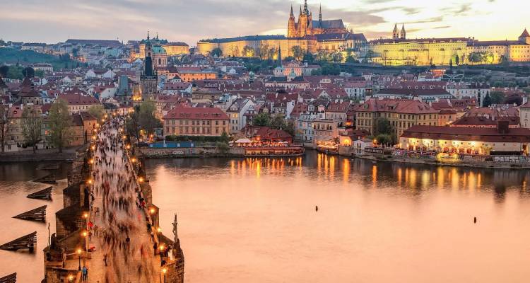 Uitzicht op Karelsbrug in Praag bij schemering, druk bezocht door wandelaars, met het verlichte kasteelheuvel op de achtergrond weerspiegeld in de rivier de Moldau.