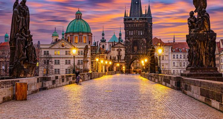 Vista de un puente histórico y edificios, con estatuas e iluminación al atardecer.