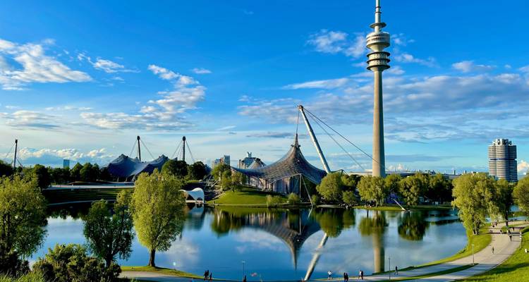 Parque verde con un lago, torre moderna y arquitectura única bajo un cielo azul.