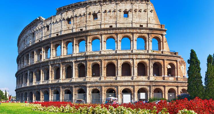 Plan large de jour du Colisée entouré de pelouse verte et de parterres de fleurs rouges, sous un ciel bleu éclatant.