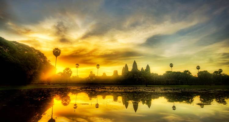 Sunrise over Angkor Wat with palm trees reflecting in a pond.