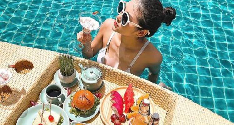Woman in pool enjoys a floating brunch tray and a drink while wearing white sunglasses.