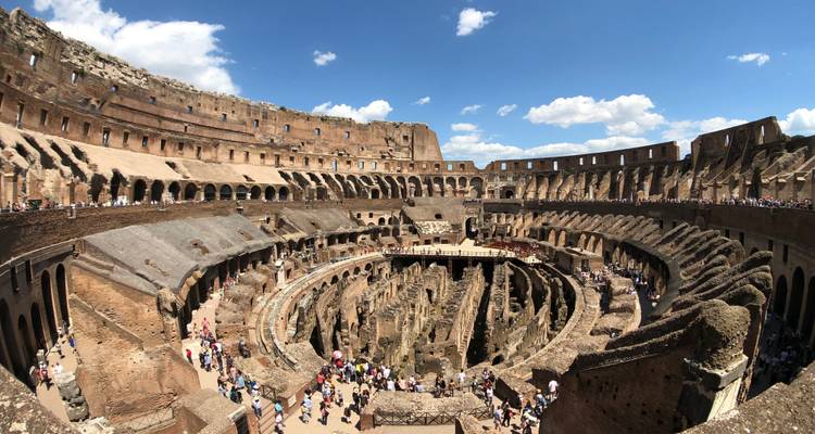 Panoramic interior view of Rome’s Colosseum filled with visitors under a blue sky.