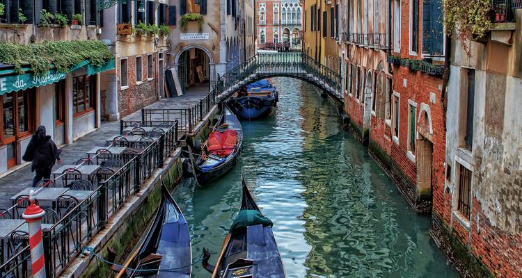 Quiet Venetian canal lined with aged brick buildings and moored gondolas beneath a small footbridge.