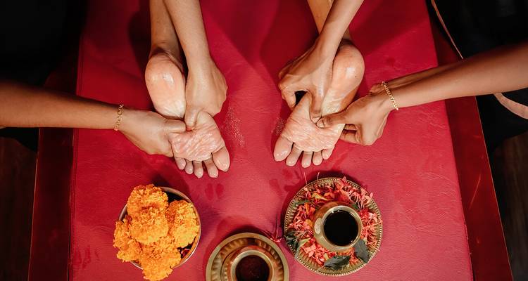 Deux mains donnant un massage des pieds sur un tapis rouge, entourées de fleurs.