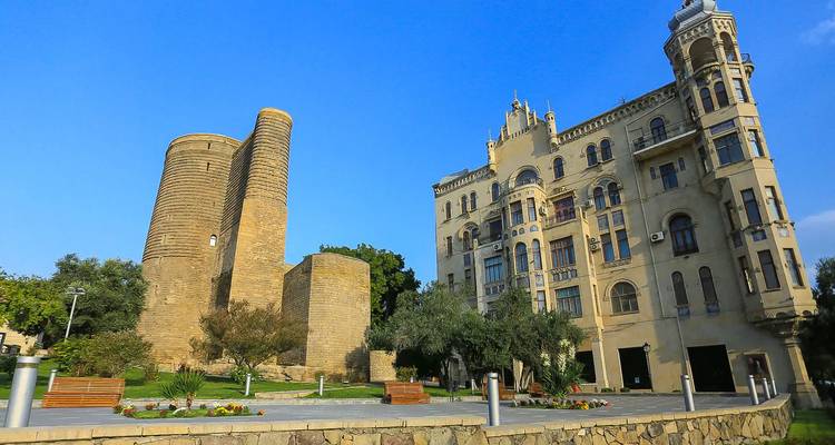 La histórica Torre de la Doncella se alza junto a un edificio ornamentado de principios del siglo XX entre jardines paisajísticos.