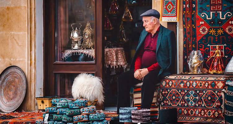 Comerciante anciano de alfombras sentado fuera de una tienda rodeado de alfombras tradicionales coloridas y artesanías.