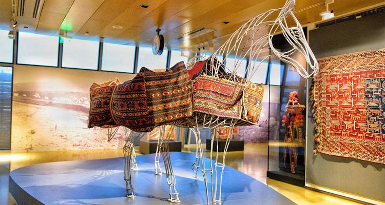 Exposition de musée intérieure présentant un cheval en structure métallique drapé de tapis azerbaïdjanais colorés, éclairé par un éclairage de galerie chaleureux.