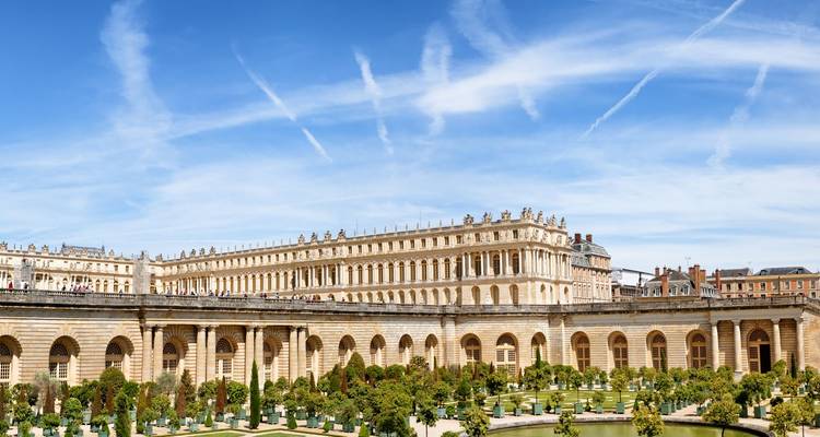 Vue panoramique de la façade du Palais de Versailles et des jardins entretenus sous un ciel bleu traversé par des traînées de condensation.