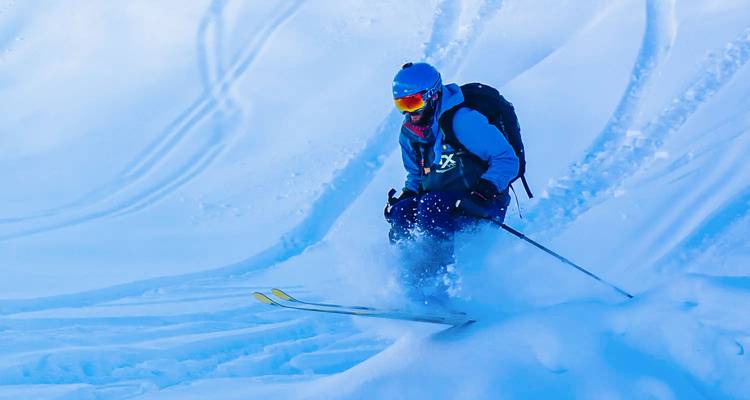 Skifahrer, der an einem strahlenden Wintertag durch frischen Pulverschnee rast und dabei eine Schneefontäne hinterlässt.