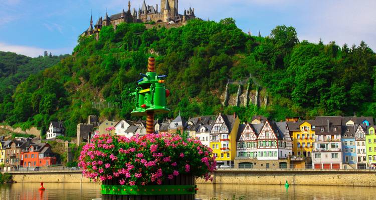 Des maisons à colombages colorées longeant la rivière Moselle avec un bac à fleurs en forme de tonneau rempli de fleurs roses et le château de Cochem perché sur la colline verdoyante au-dessus.
