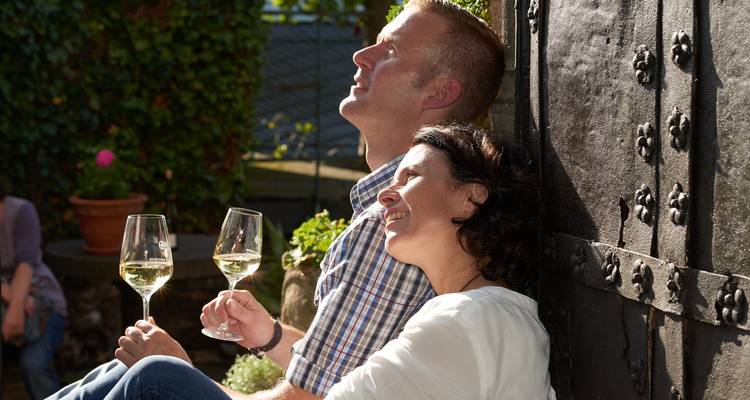 Couple souriant se détend contre une vieille porte clouée en fer, savourant des verres de vin blanc à la lumière du soleil chaud.