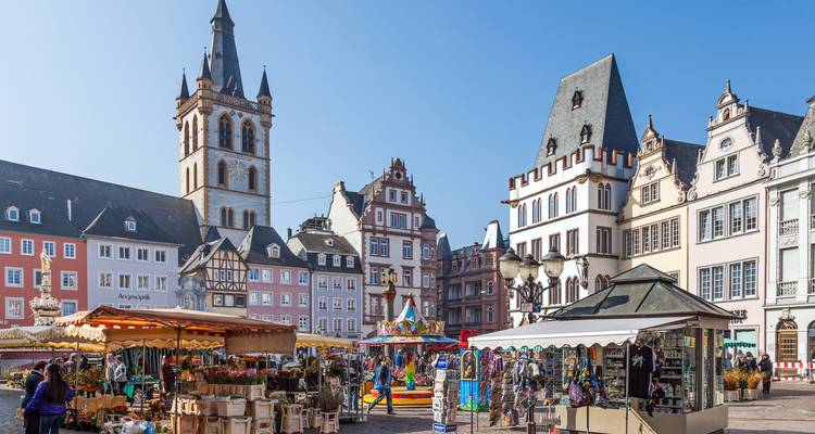 Place du marché animée à Trèves avec des bâtiments historiques aux couleurs pastel et un haut clocher d'église sous un ciel bleu dégagé.