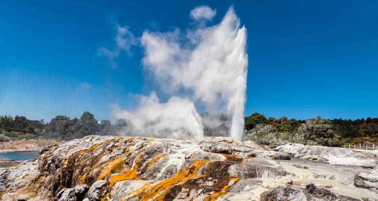 Un geyser puissant en éruption, jaillissant haut dans un ciel bleu clair au-dessus de terrasses minérales colorées.