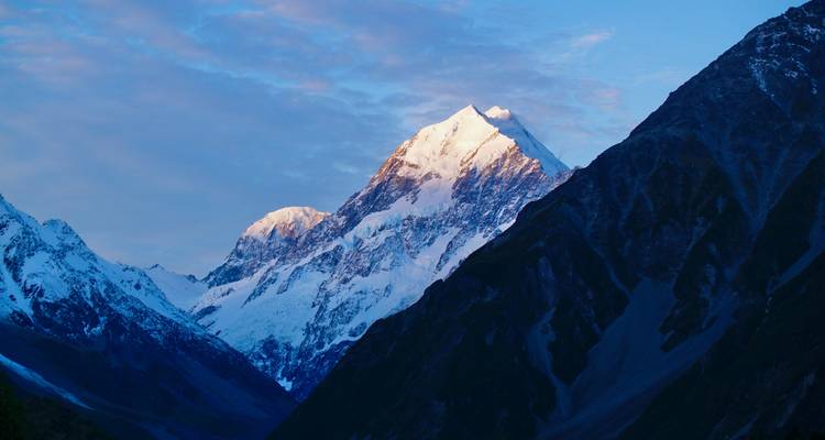 Pic enneigé du Mont Cook brillant au coucher du soleil contre un ciel bleu doux surplombant les versants de la vallée sombre.