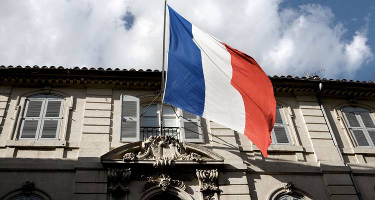 Drapeau tricolore français flottant au-dessus d'un bâtiment en pierre classique contre un ciel partiellement nuageux.