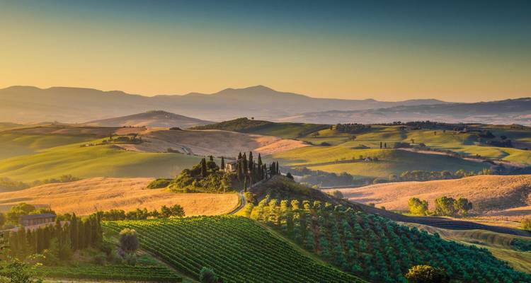 La luz del atardecer baña las colinas ondulantes clásicas y las casas de campo de la Toscana en tonos cálidos.