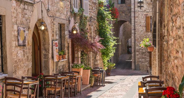 Encantador callejón de piedra estrecho con mesas de café al aire libre y plantas en flor en un pueblo medieval.