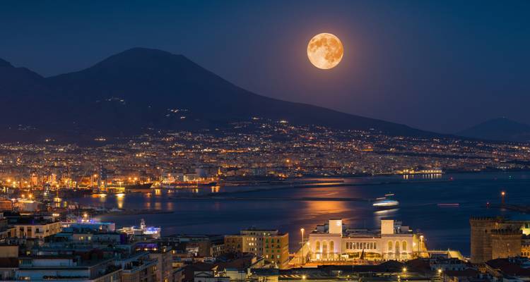 Pleine lune se levant au-dessus de la baie illuminée de Naples avec le mont Vésuve en silhouette contre le ciel nocturne.