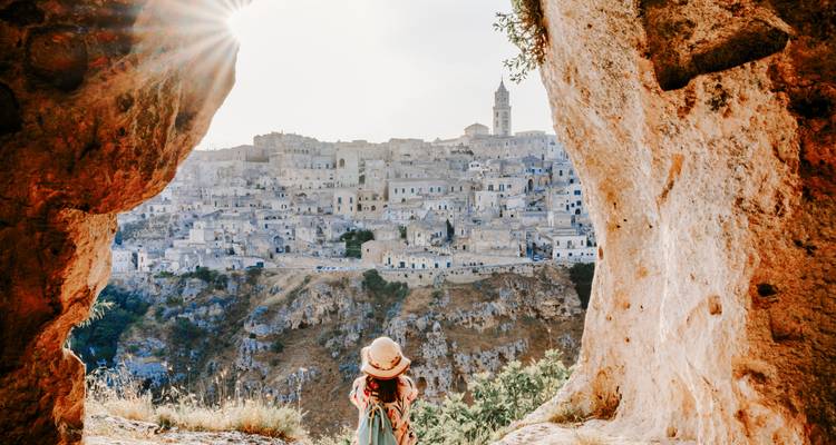 Une voyageuse portant un chapeau de soleil se tient à l'entrée d'une grotte et contemple la ville de pierre de Matera baignée dans la lumière matinale.