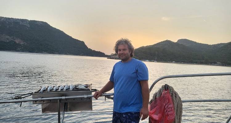 Persoon die op een boot staat met bergen op de achtergrond, vis aan het grillen.