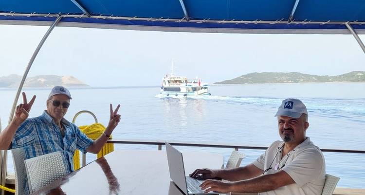 Twee mannen op een bootdek met een laptop; veerboot op de achtergrond.
