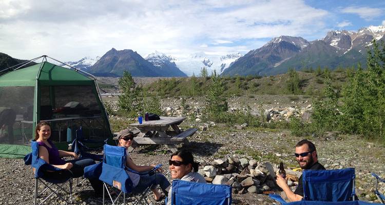 Groupe de campeurs se détendant dans des chaises pliantes à côté d'une tente avec des montagnes enneigées en arrière-plan.