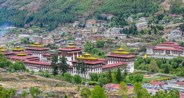 Panoramic view of an expansive white-walled dzong complex with red roofs set amid a forested valley town.